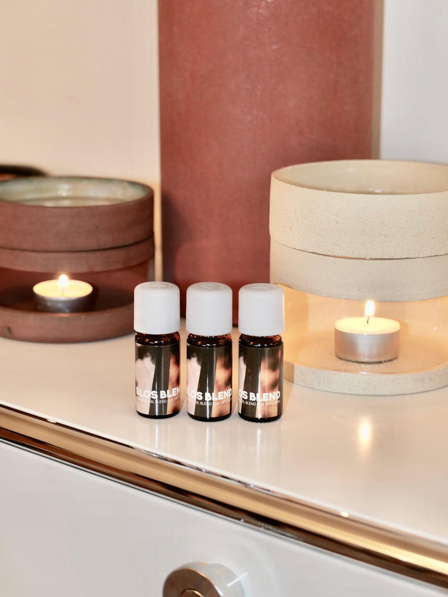Three small bottles Blos Blend with white caps on a reflective surface with oil diffusersin the background.