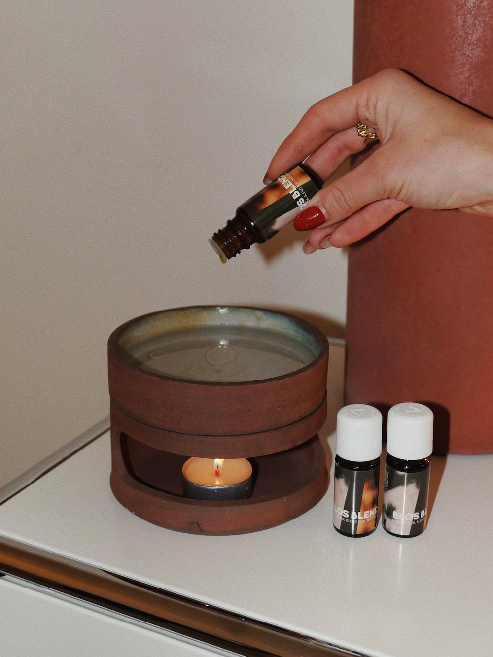Hand holding an essential oil bottle above a ceramic diffuser with candles on a surface and and essential oil blend by blos