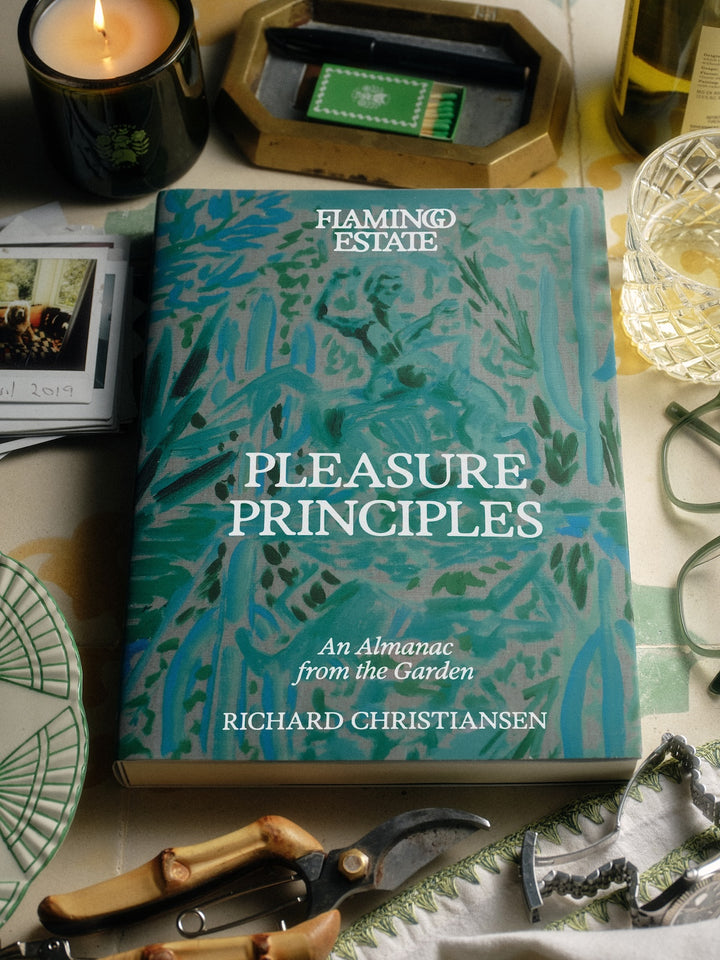 A book titled "Pleasure Principles: An Almanac from the Garden" by Richard Christiansen, placed on a tabletop surrounded by garden and kitchen props including pruning shears, a candle, glasses, and a plate.