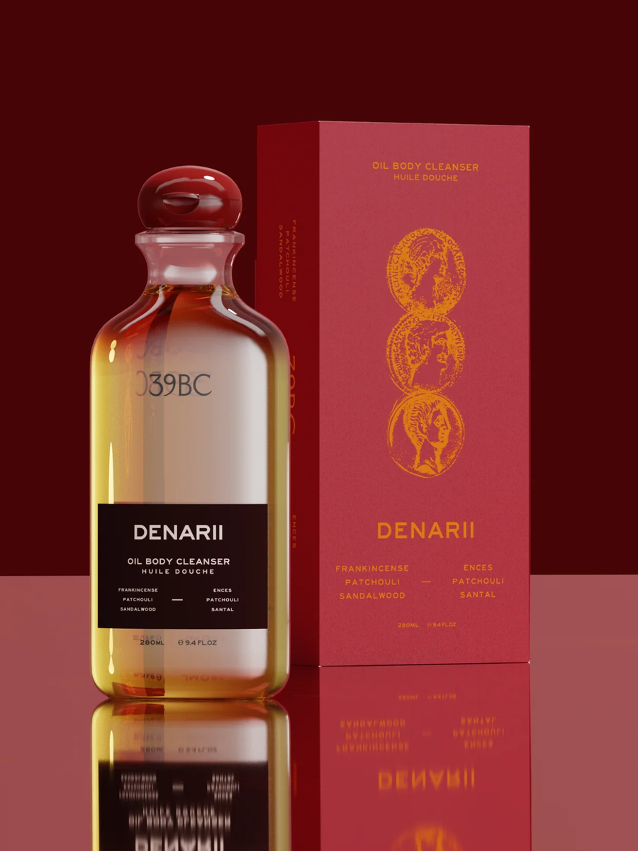 A bottle of DENARII Oil Body Cleanser with amber liquid and a black label, standing next to its red packaging box with gold lettering, on a reflective dark red surface.