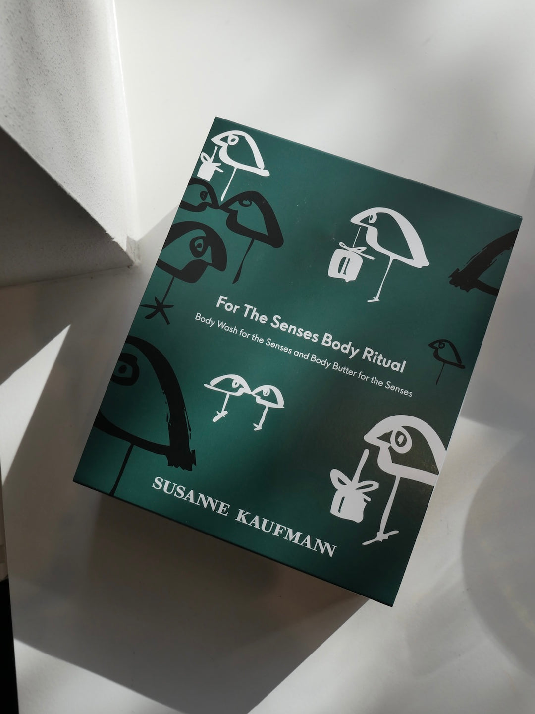 A square product box for 'For The Senses Body Ritual' by Susanne Kaufmann, featuring a dark teal-green cover with white line drawings of umbrellas and body parts, photographed on a white surface with dramatic shadows.