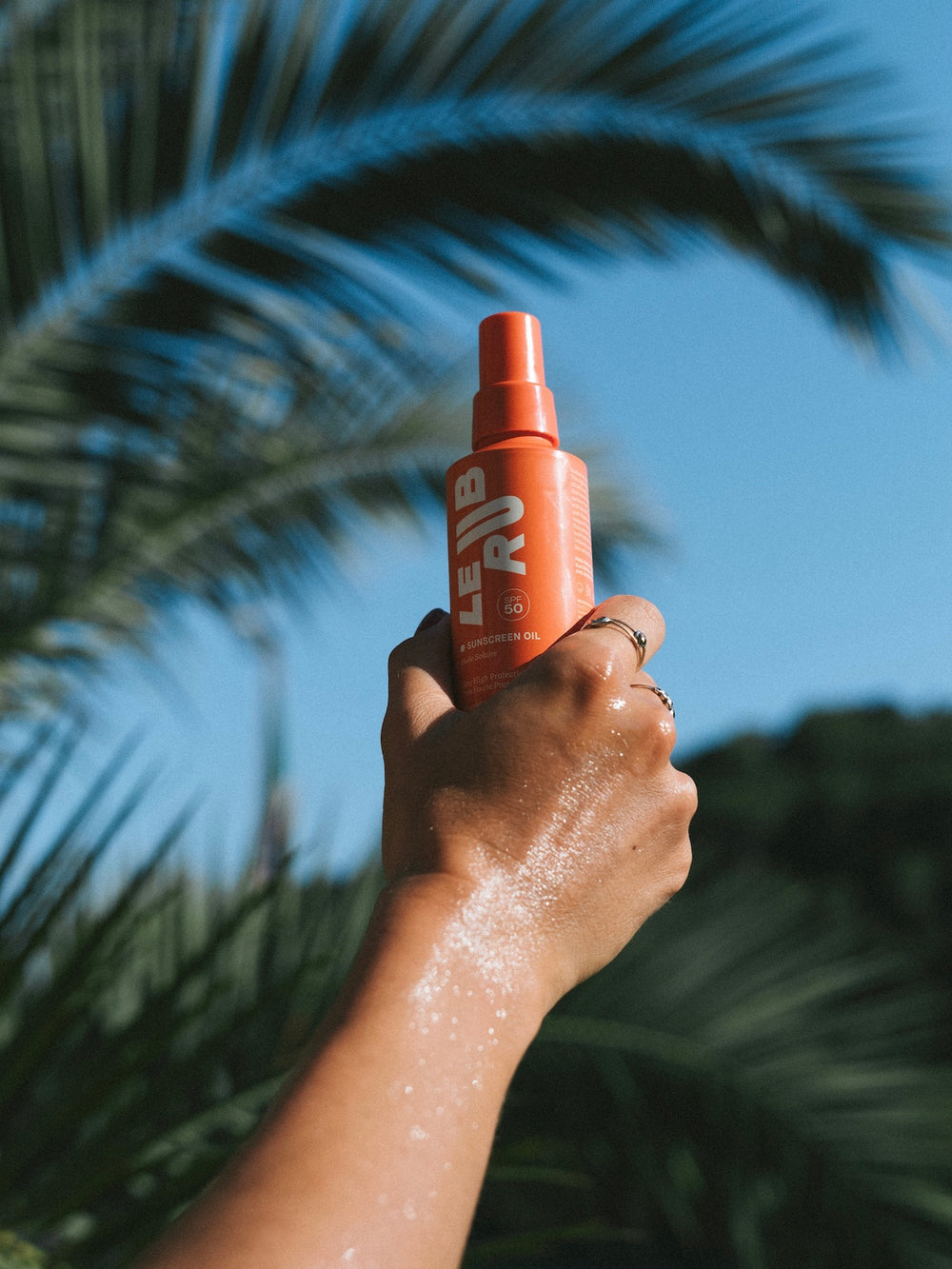 Hand holding an orange spray bottle labeled Le Rub Sunscreen Oil SPF50 against a palm tree background in Italy