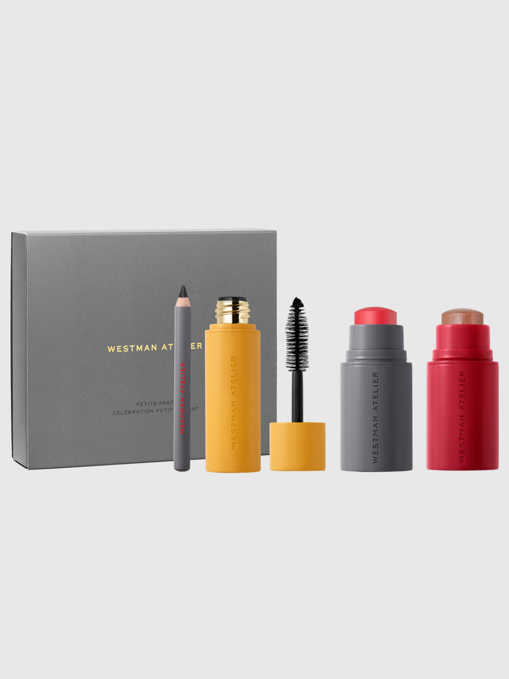 Westman Atelier makeup set featuring a gray box, a gray pencil, a yellow mascara tube with wand, a gray lip product with red cap, and a red lip product with beige cap, arranged on a light gray background.