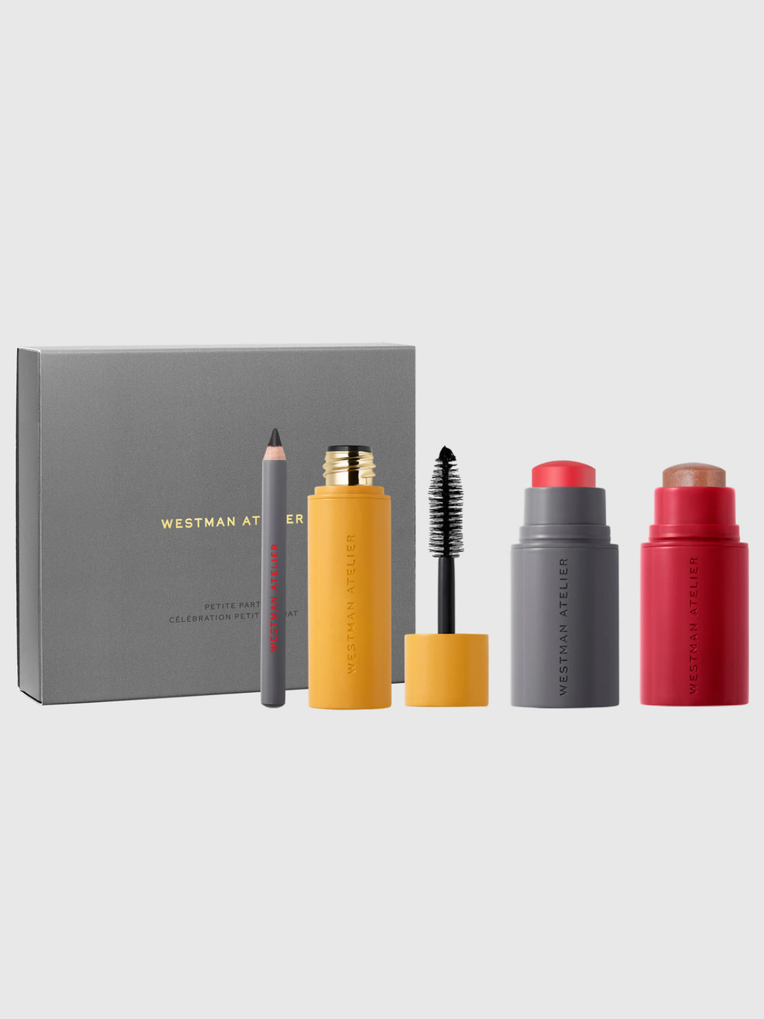 Westman Atelier makeup set featuring a gray box, a gray pencil, a yellow mascara tube with wand, a gray lip product with red cap, and a red lip product with beige cap, arranged on a light gray background.