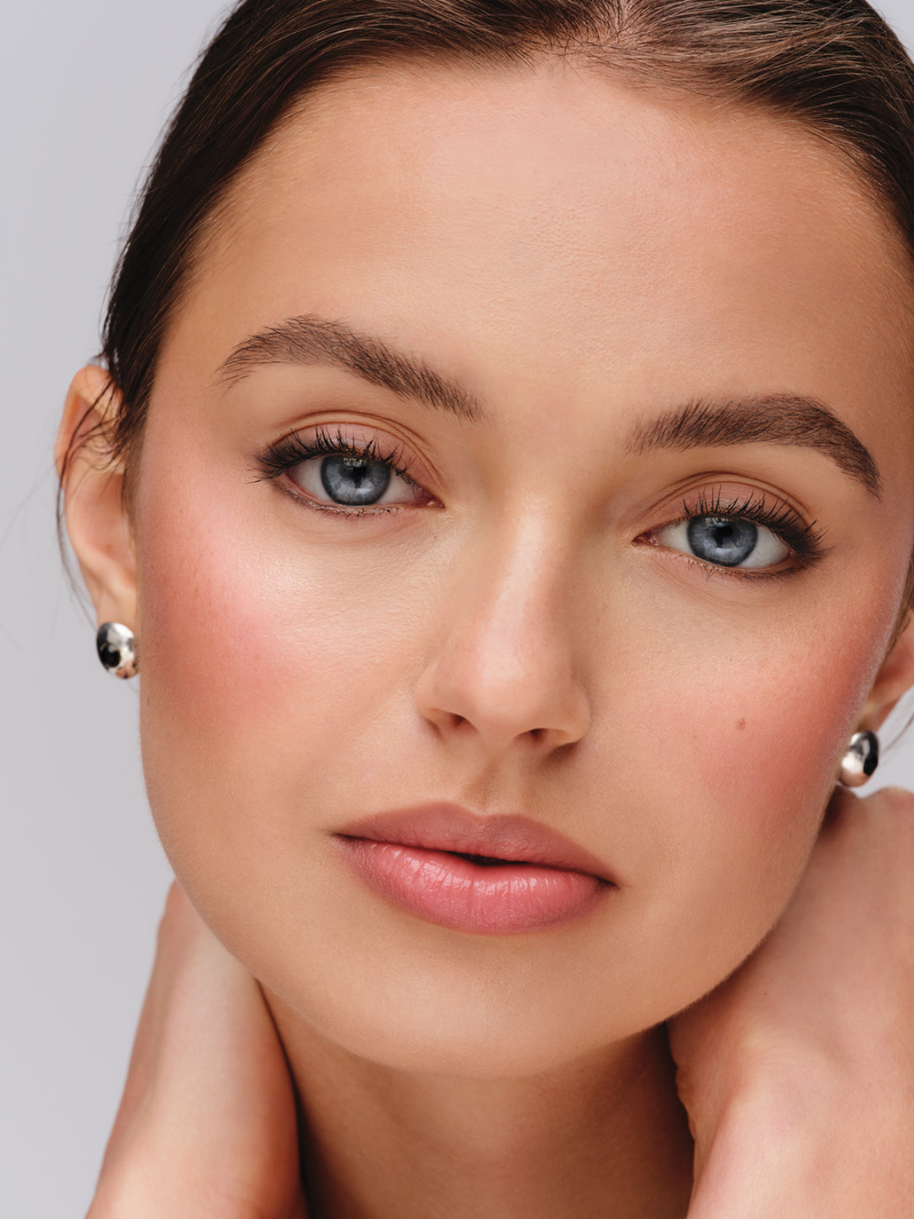 A close-up portrait of a woman with blue eyes, wearing small silver stud earrings, against a light gray background.