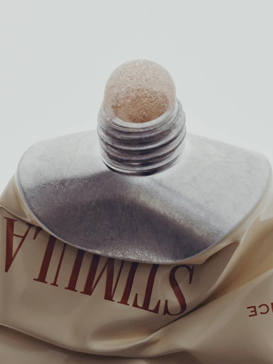 A close-up of a metallic silver squeeze tube with a textured beige cap, resting on draped cream-colored fabric printed with the word 'STIMULA'.