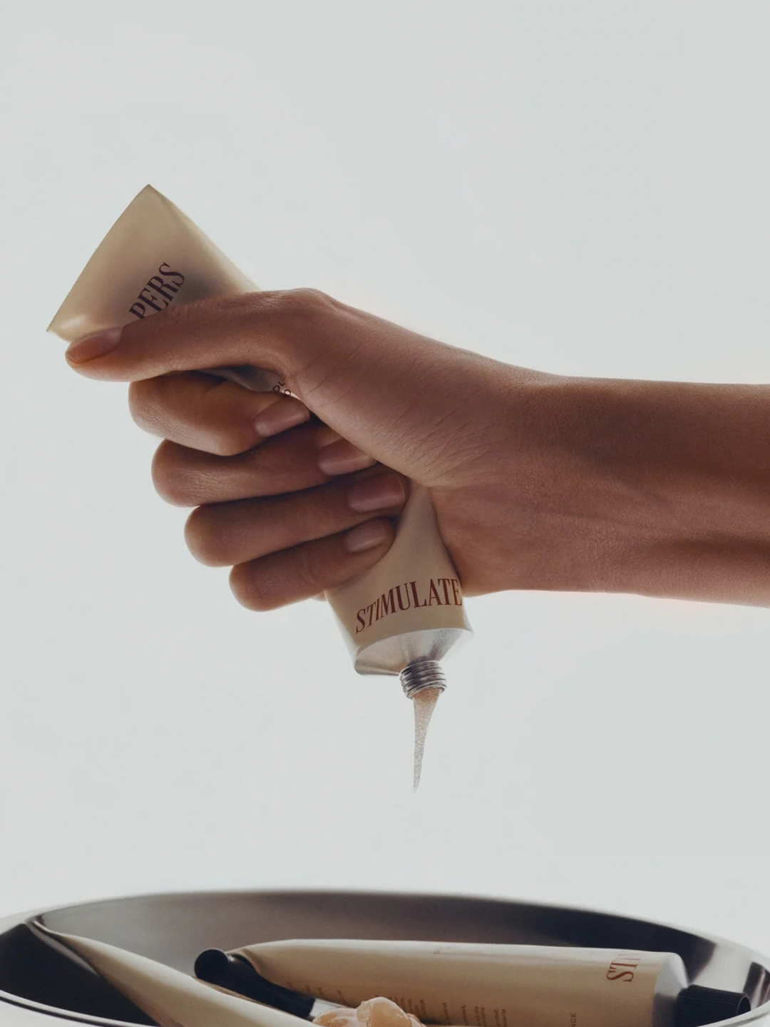 A hand squeezes a beige cosmetic tube, with cream dripping from the nozzle, over a tray holding other similar tubes. The background is a clean, bright off-white studio backdrop.