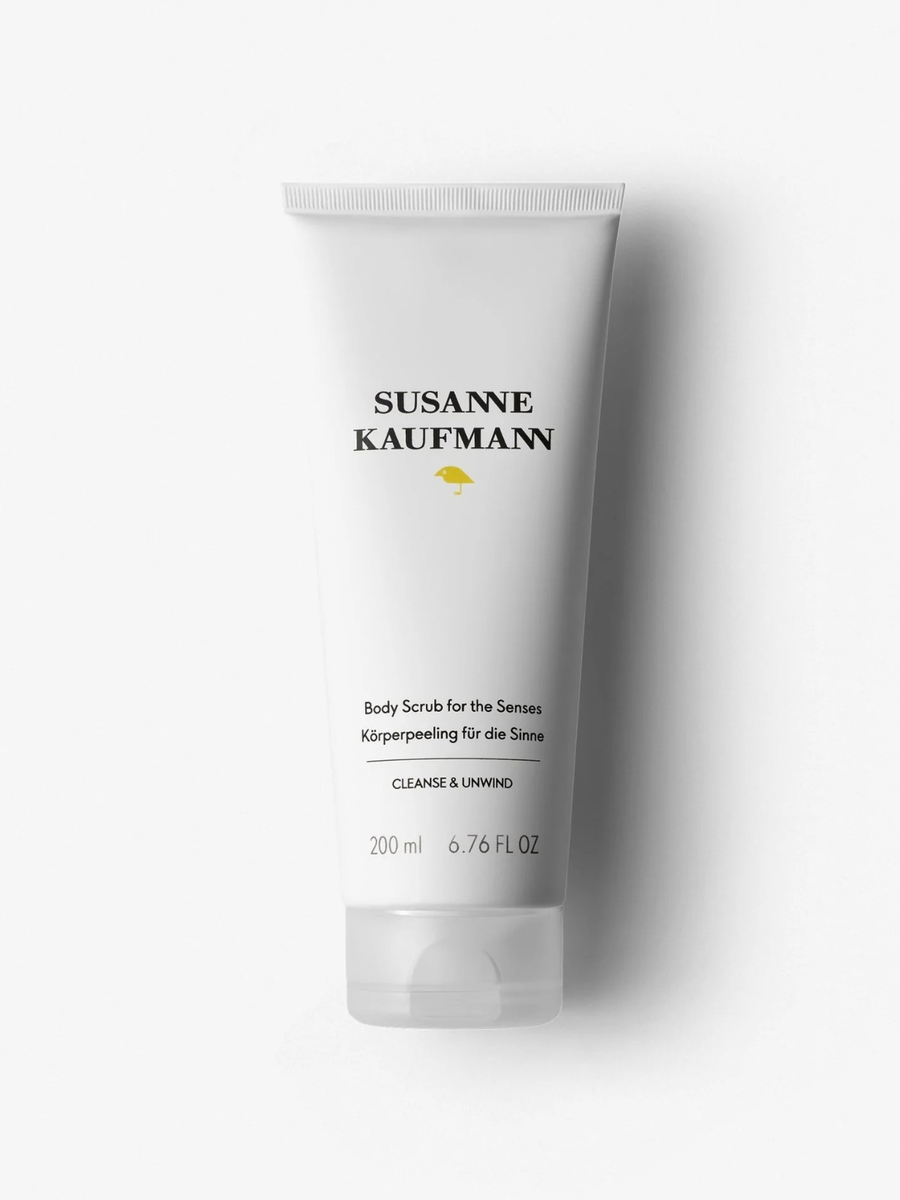 A white squeeze tube of Susanne Kaufmann body scrub with black text and a small yellow umbrella logo, photographed on a clean white background with a soft shadow.