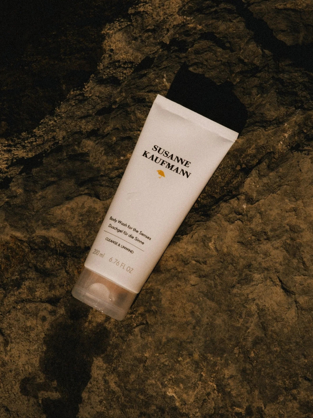 A white tube of Susanne Kauffmann body wash for the sensitive skin, lying diagonally on a dark, textured rock surface.