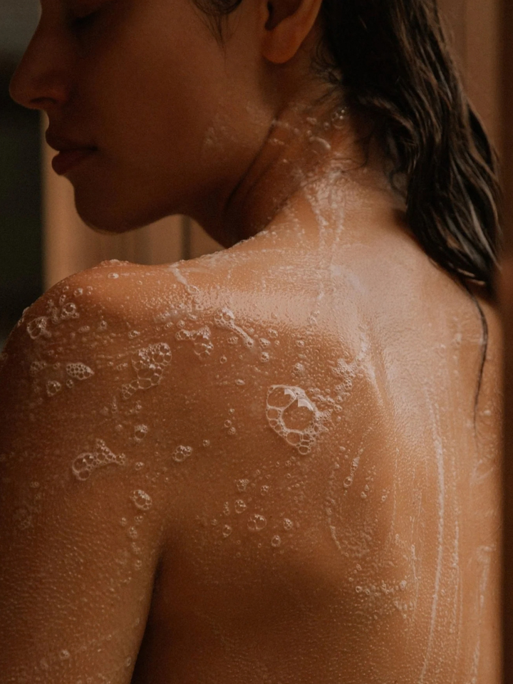 Close-up of a person's bare shoulder and upper back covered in white soap lather and bubbles, with wet dark hair visible against a warm brown-toned background.