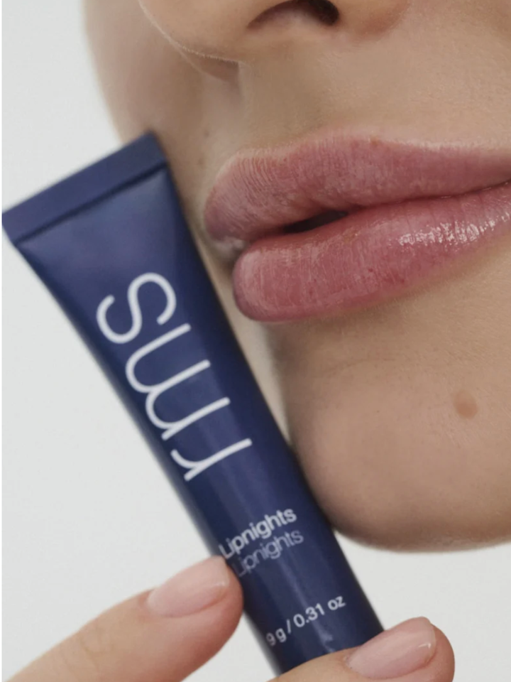 Blue lip balm tube with 'rms' branding held by a hand, close-up of lips in the background.